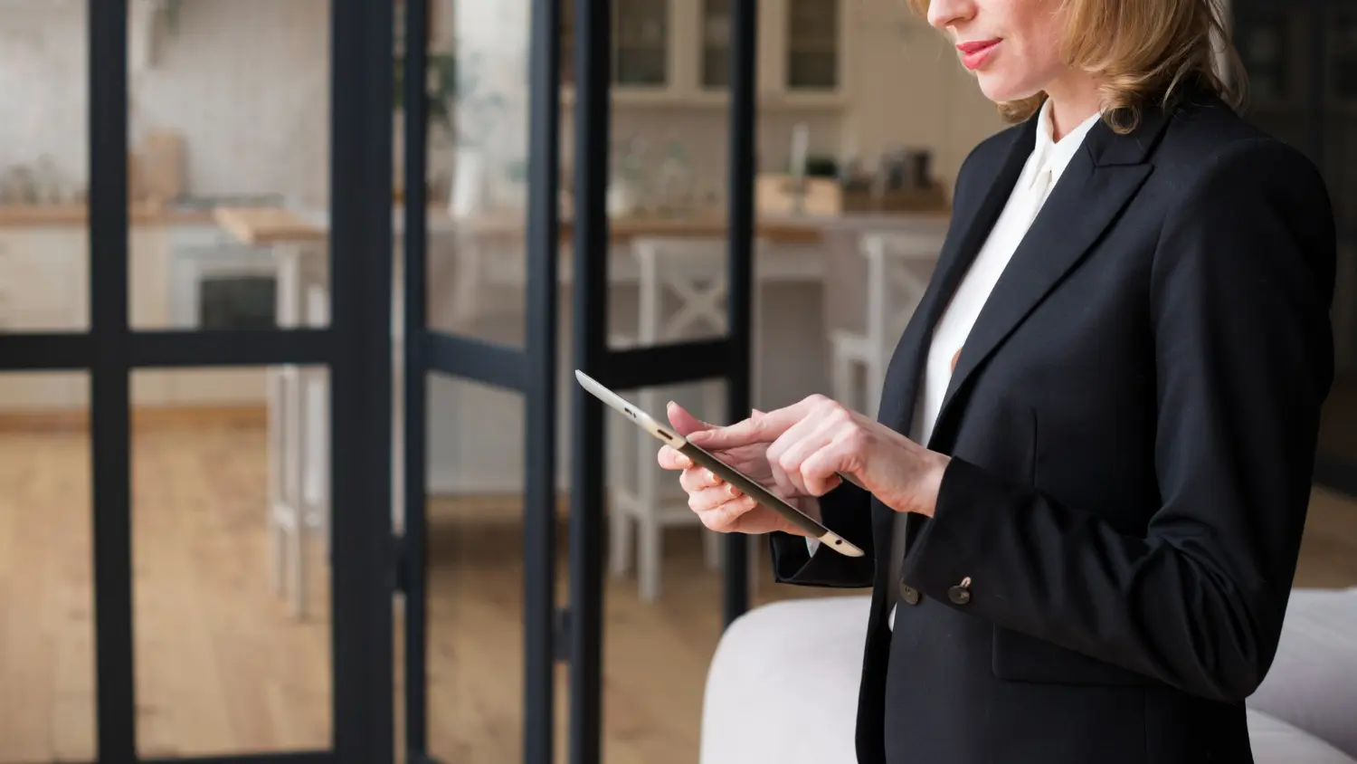 Une femme en tenue professionnelle utilise une tablette numérique dans un intérieur moderne, près d’une cloison vitrée.