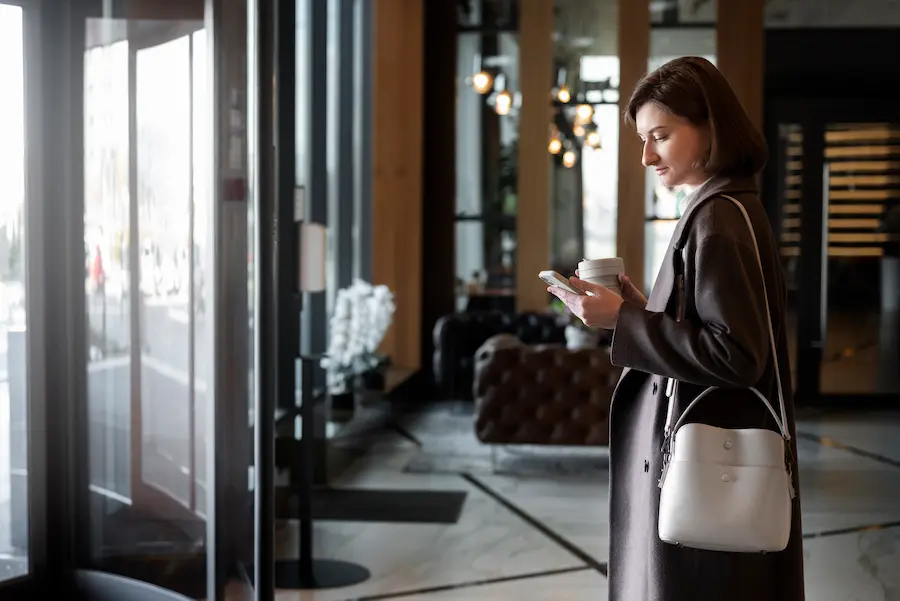 Woman in a hotel lobby checking her smartphone while holding a coffee cup, illustrating the use of a digital service for hotel guests.