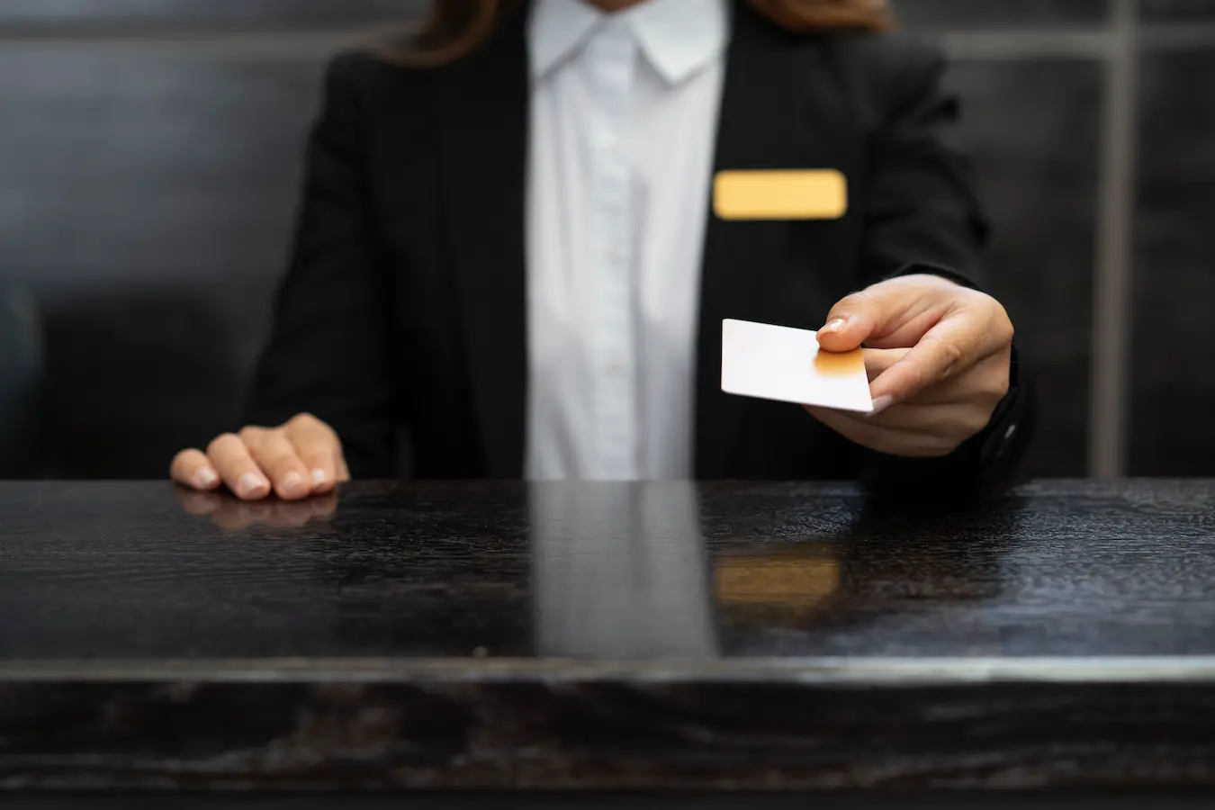 Hotel receptionist in professional attire handing over a key card above a black marble front desk counter, symbolizing check-in or guest arrival.