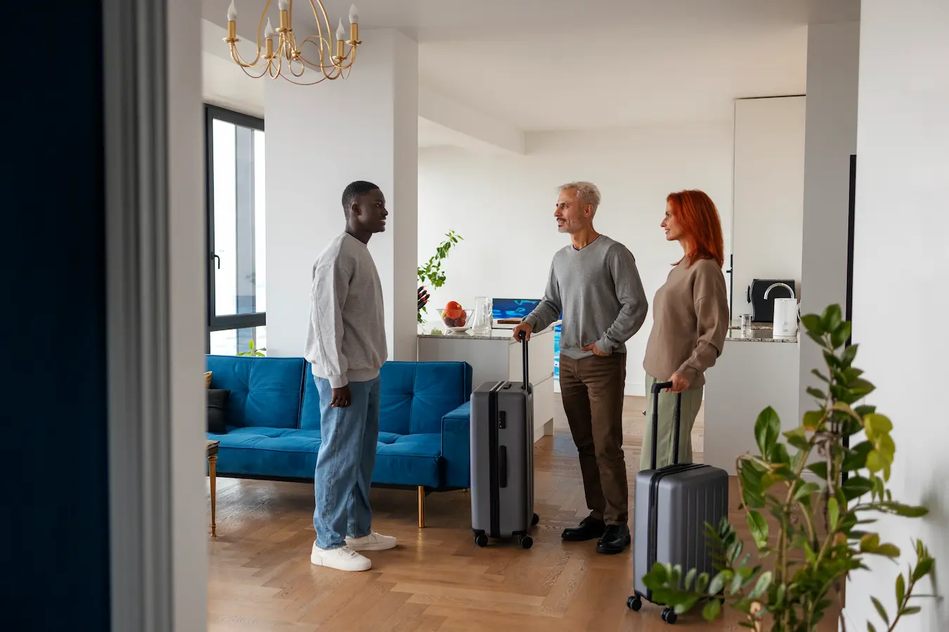 Un hôte souriant accueille un couple de voyageurs avec leurs valises dans un appartement moderne et lumineux.