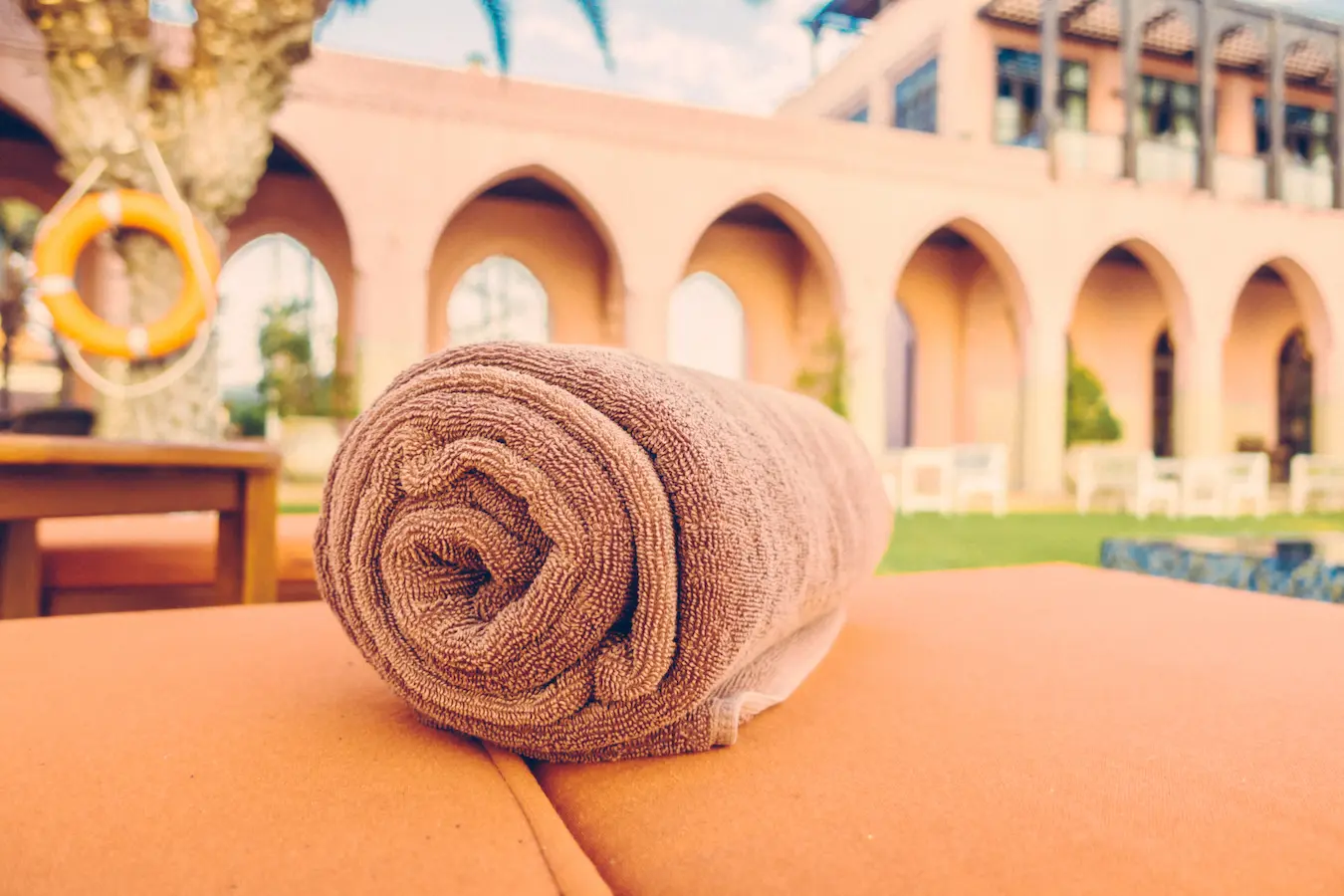 Rolled brown towel placed on a sun lounger near a pool, with a Mediterranean-style building featuring arches in the background.