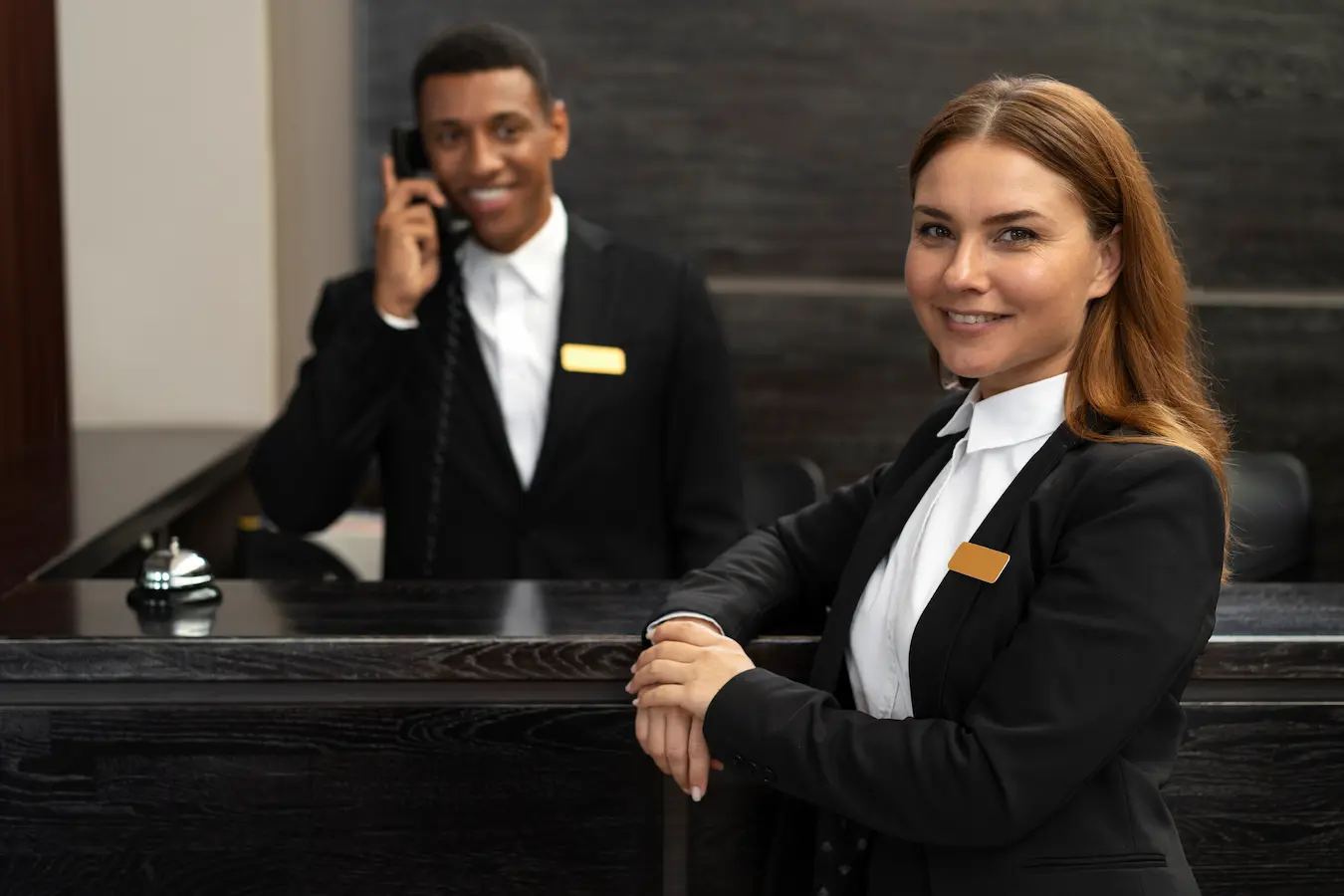 Two hotel receptionists in formal uniforms assisting guests at a front desk, with one smiling at the camera and the other speaking on the phone.