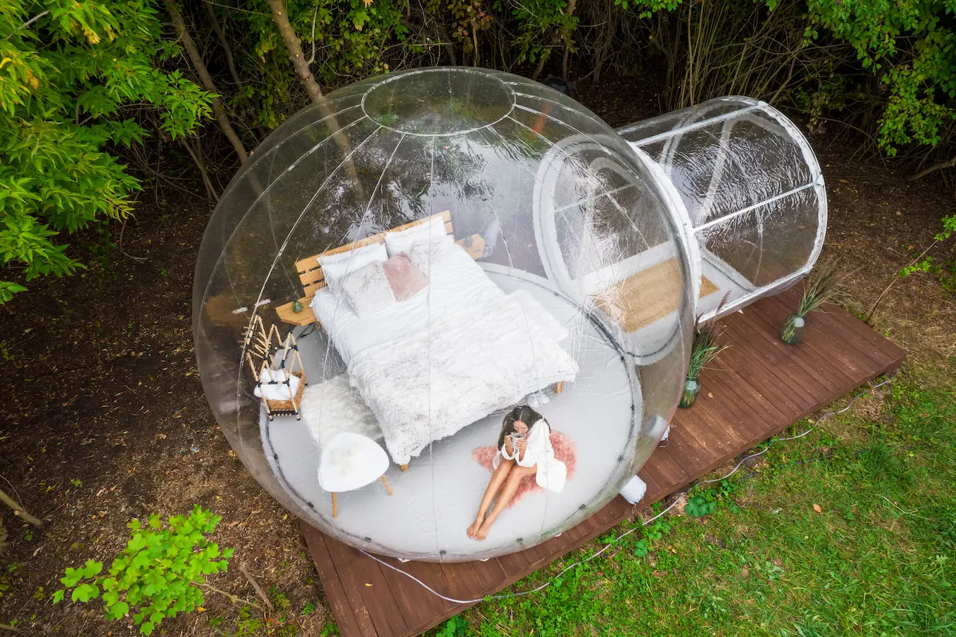 Aerial view of a woman relaxing inside a transparent bubble tent with a bed, set in the middle of nature.