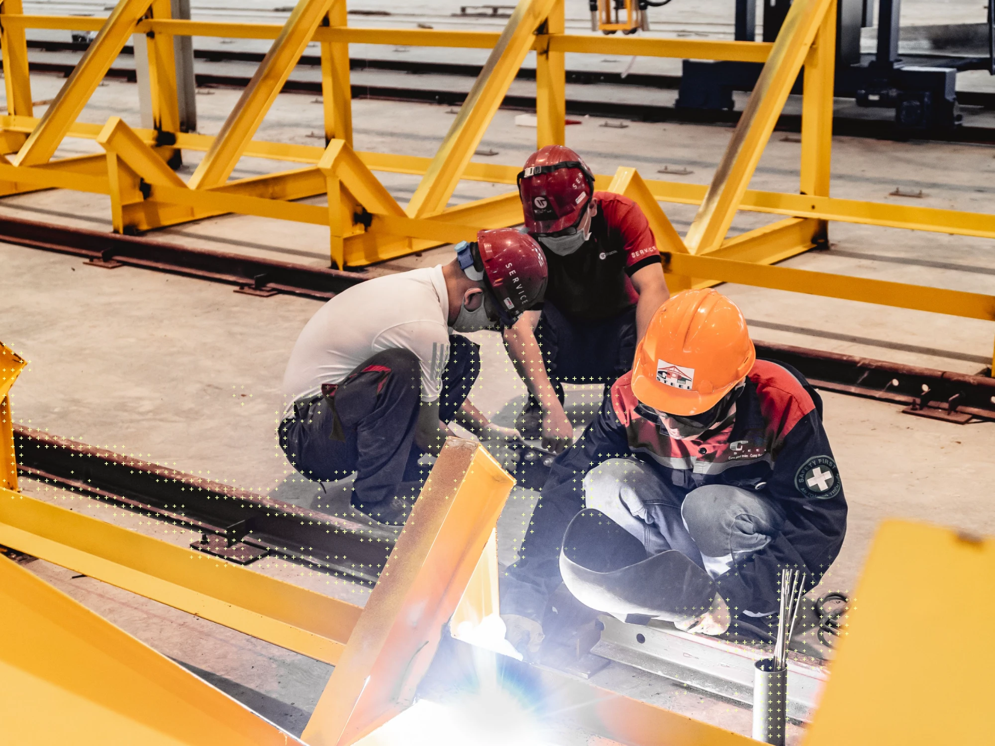 Three workers in hard hats welding a steel frame, with a data-dot overlay