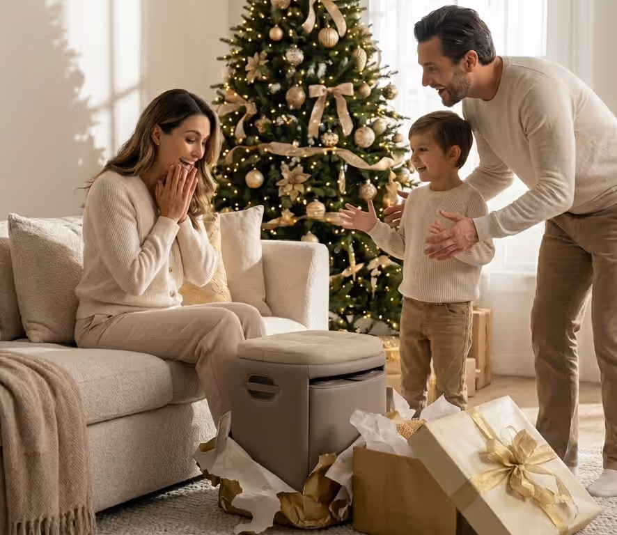 Une femme surprise assise sur un canapé, un homme et un enfant souriants près d'un sapin de Noël décoré, entourés de cadeaux déballés.