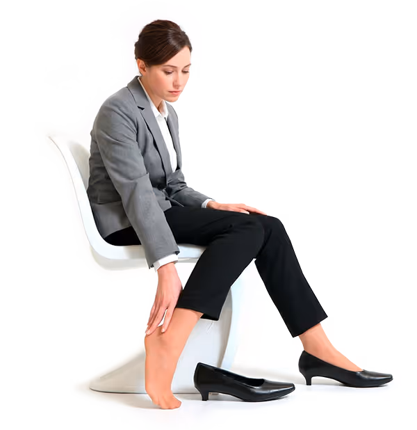 Femme en costume assise sur une chaise, retirant une chaussure noire et massant son pied gauche, semblant avoir mal aux pieds.