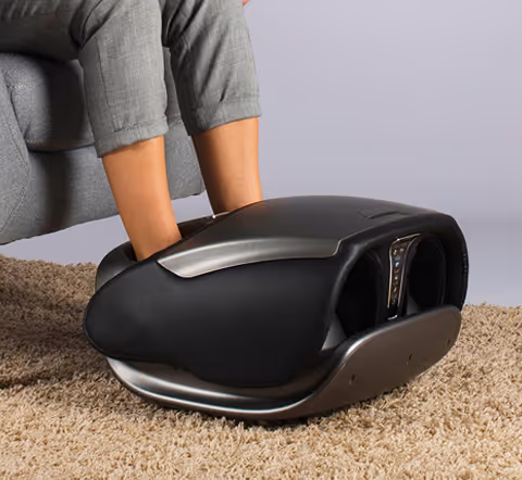 Personne assise utilisant un appareil de massage des pieds Pedimass noir sur un tapis beige.