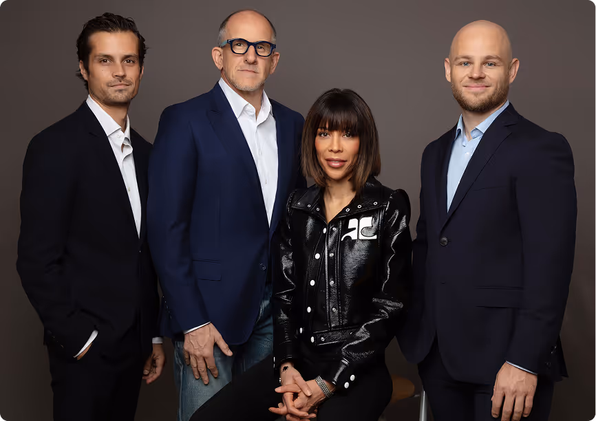 Quatre professionnels en tenue formelle posant devant un fond gris, dont une femme assise en veste noire brillante et trois hommes debout en vestons sombres.