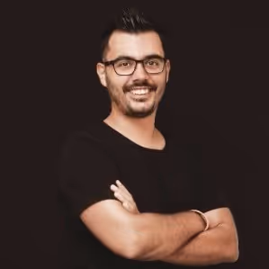 Esteve Mata sonríe, con gafas y barba, vestido con una camiseta negra y los brazos cruzados frente a un fondo oscuro.