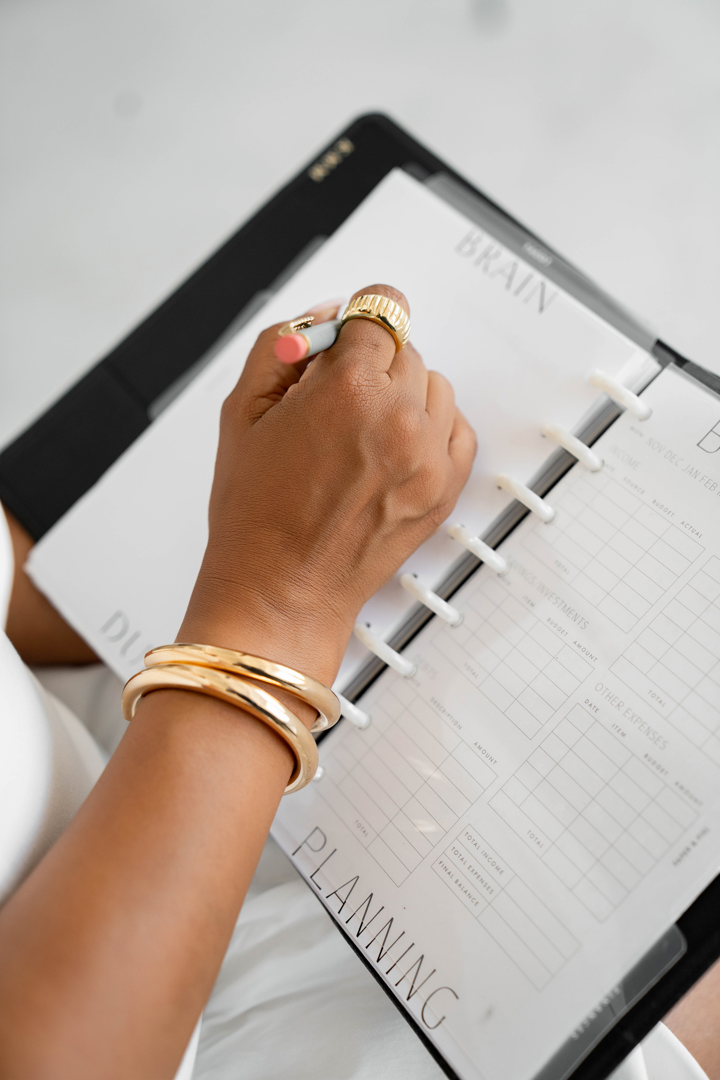 Person with gold bracelets and rings writing in a financial planning organizer with categories for income, savings, investments, and expenses.