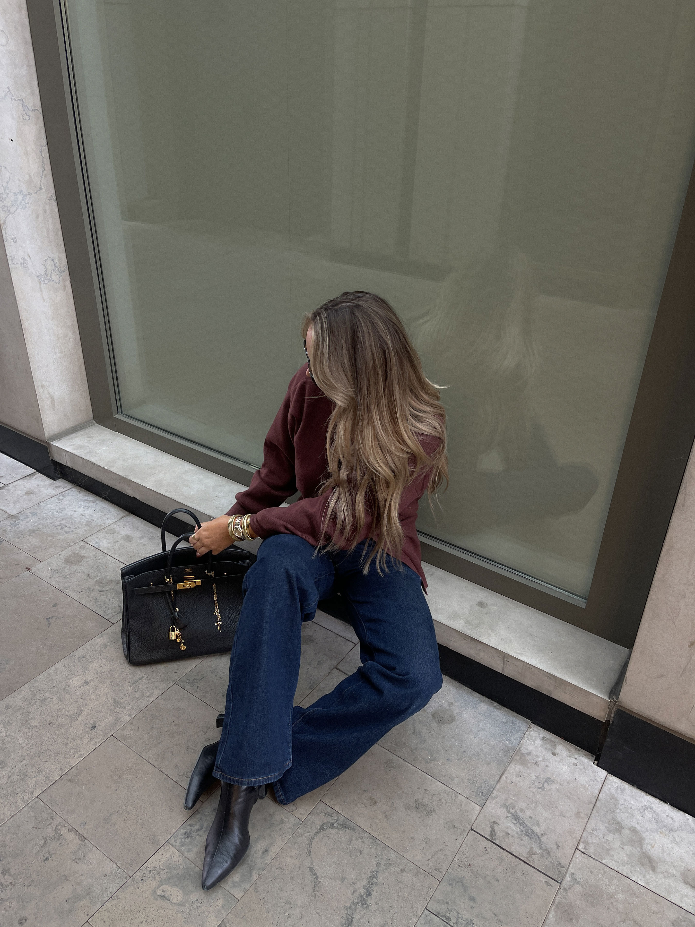Woman with long hair sitting on a stone floor against a window, wearing a burgundy sweater, blue jeans, black pointed boots, and holding a black handbag.