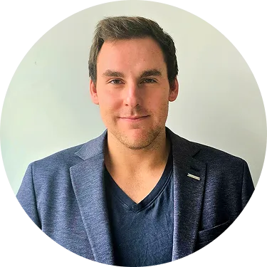 Porträt eines lächelnden Mannes mit kurzem braunem Haar, der einen dunkelblauen Blazer und ein schwarzes T-Shirt trägt, vor hellem Hintergrund.