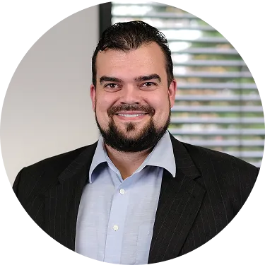 Lächelnder Mann mit dunklem Haar, Bart und Anzug vor einem Fenster mit Jalousien.