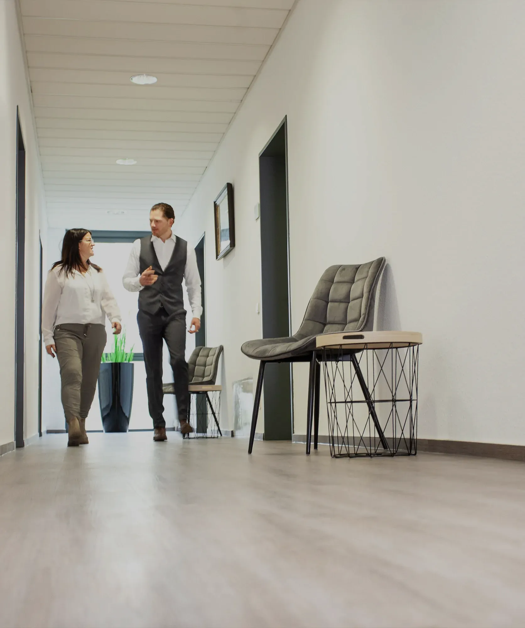 Zwei Personen, die in einem hellen Flur mit grauen Stühlen und kleinen Tischen auf der rechten Seite spazieren und sich unterhalten.