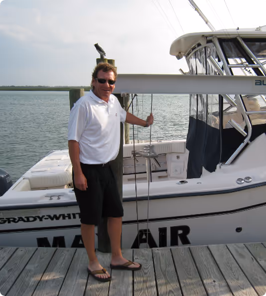 Man wearing sunglasses, white polo shirt, black shorts, and flip-flops standing on a wooden dock next to a Grady-White boat named Mare Air on calm water.