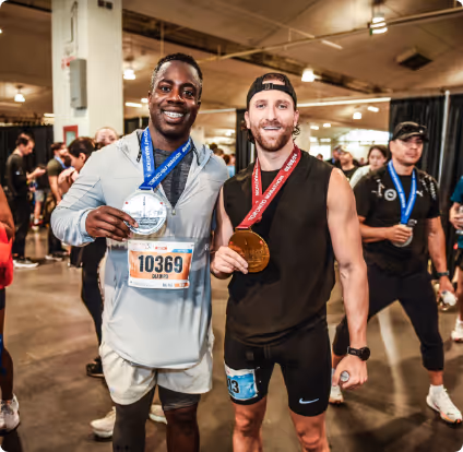 Two male runners smiling and holding medals indoors after a race, with other participants in the background.