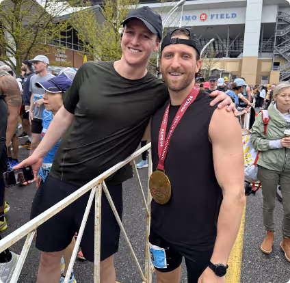 Two men smiling and posing together at an outdoor event, one wearing a finisher medal and race bib.