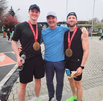 Two men wearing black athletic clothing and medals stand on either side of a man in a light blue shirt and gray cap, all smiling outdoors on a paved area.