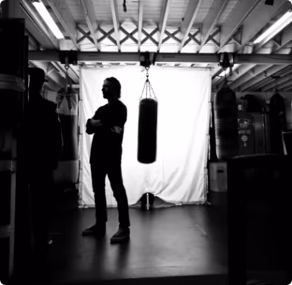 Silhouette of a person standing with arms crossed near a hanging punching bag in a gym with exposed beams.