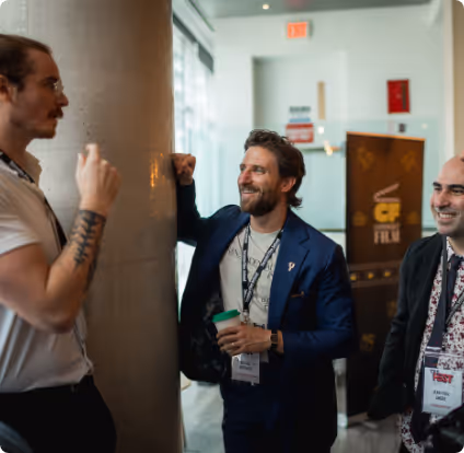 Three men smiling and chatting in a conference hallway, one wearing a blue blazer and holding a cup.