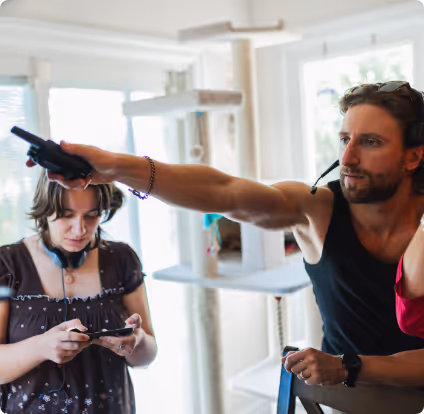 A man aiming a gaming gun controller while a woman looks at her phone headset around her neck in a bright room.