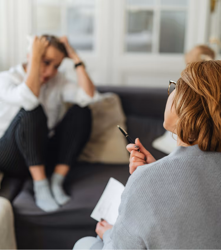 Therapist taking notes while client sits with head in hands on couch during therapy session.