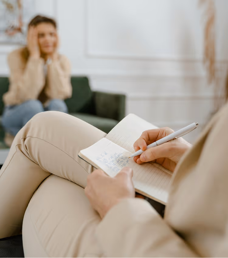 Therapist writing notes while patient sits in green chair in background during counseling session.
