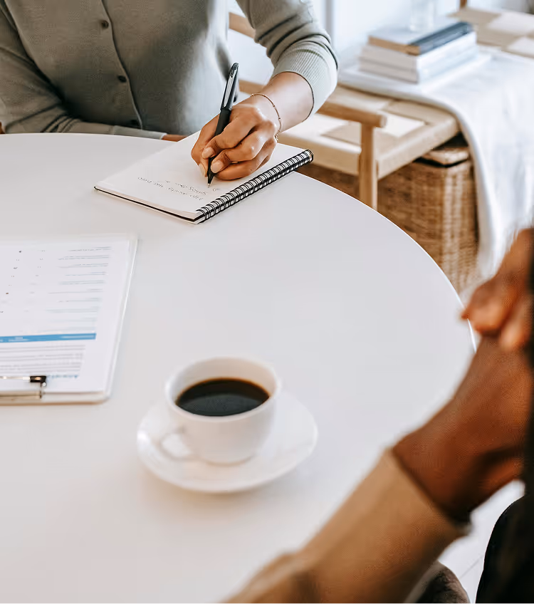 Person writing in notebook at white table with coffee cup, another person's hands visible across the table.