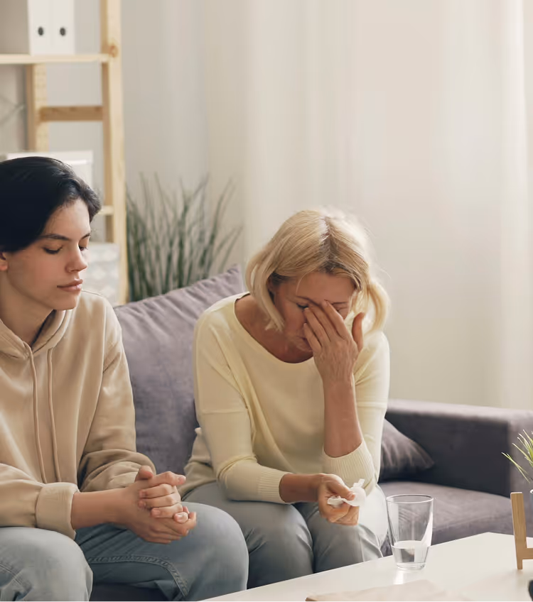 Two women sitting on a couch, one with her head in her hands appearing distressed while the other sits beside her looking concerned.