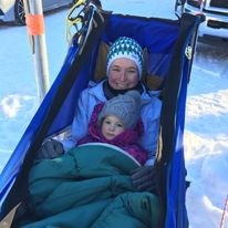 Mom and child bundled in winter clothes sitting together in a blue sled on snow.