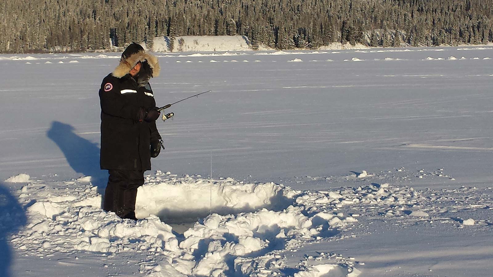 Person in a black parka ice fishing on a frozen lake with a snow-covered forest in the background.