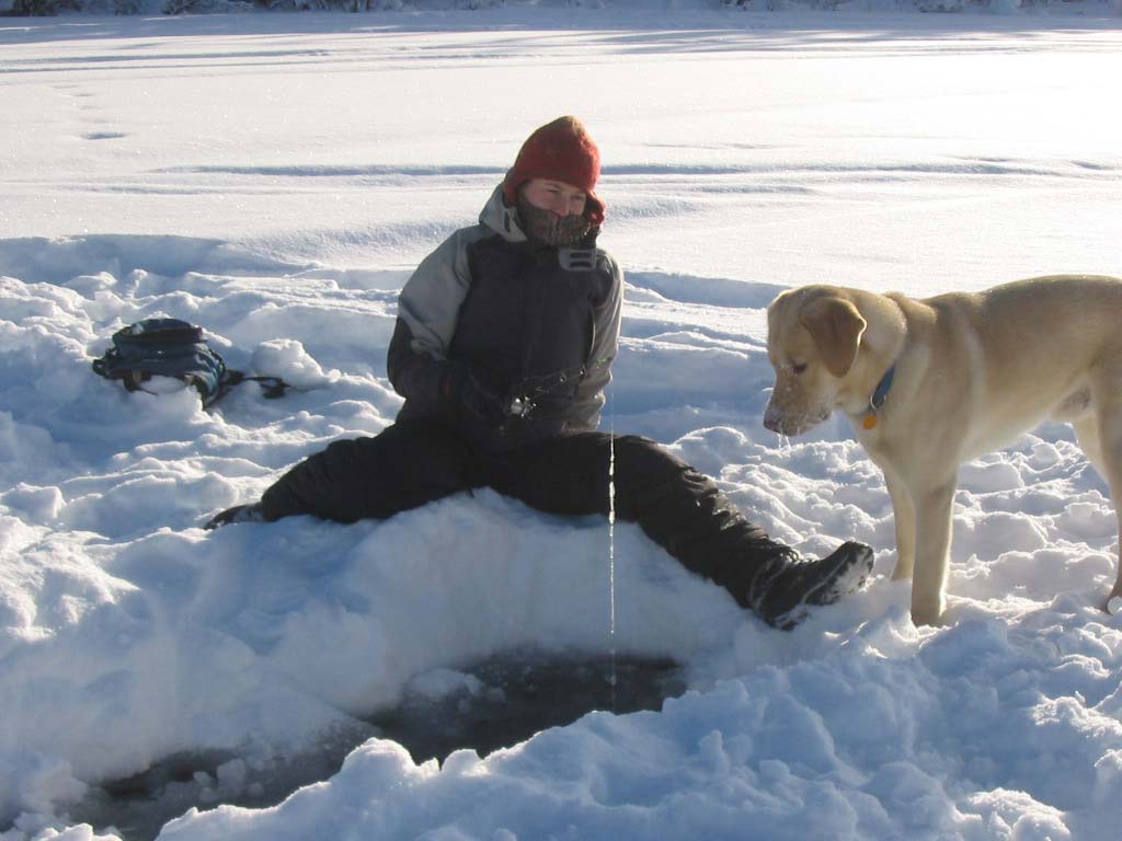 Person sitting on snow with legs spread, ice fishing through hole, watched by a yellow Labrador dog.
