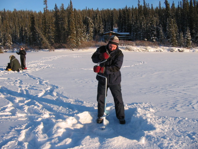 Person wearing winter clothes using an ice auger to drill a hole on a frozen lake surrounded by snowy forest.