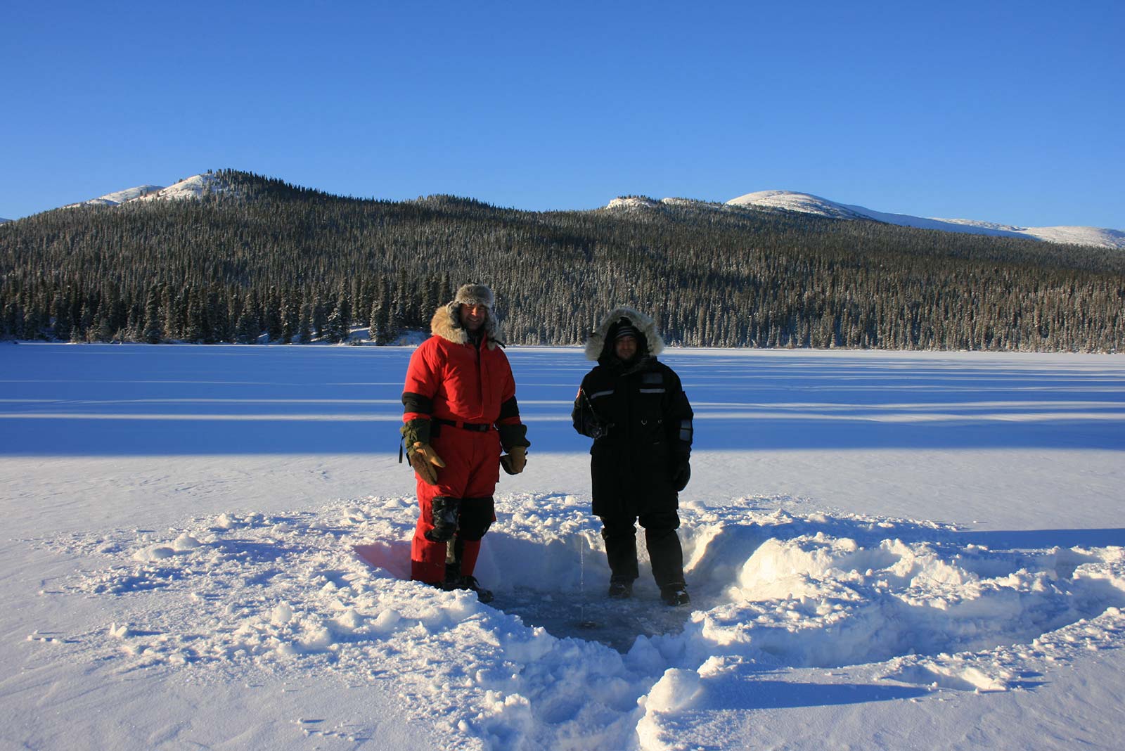 Two people in winter gear standing on a frozen lake with a hole in the ice surrounded by snow.