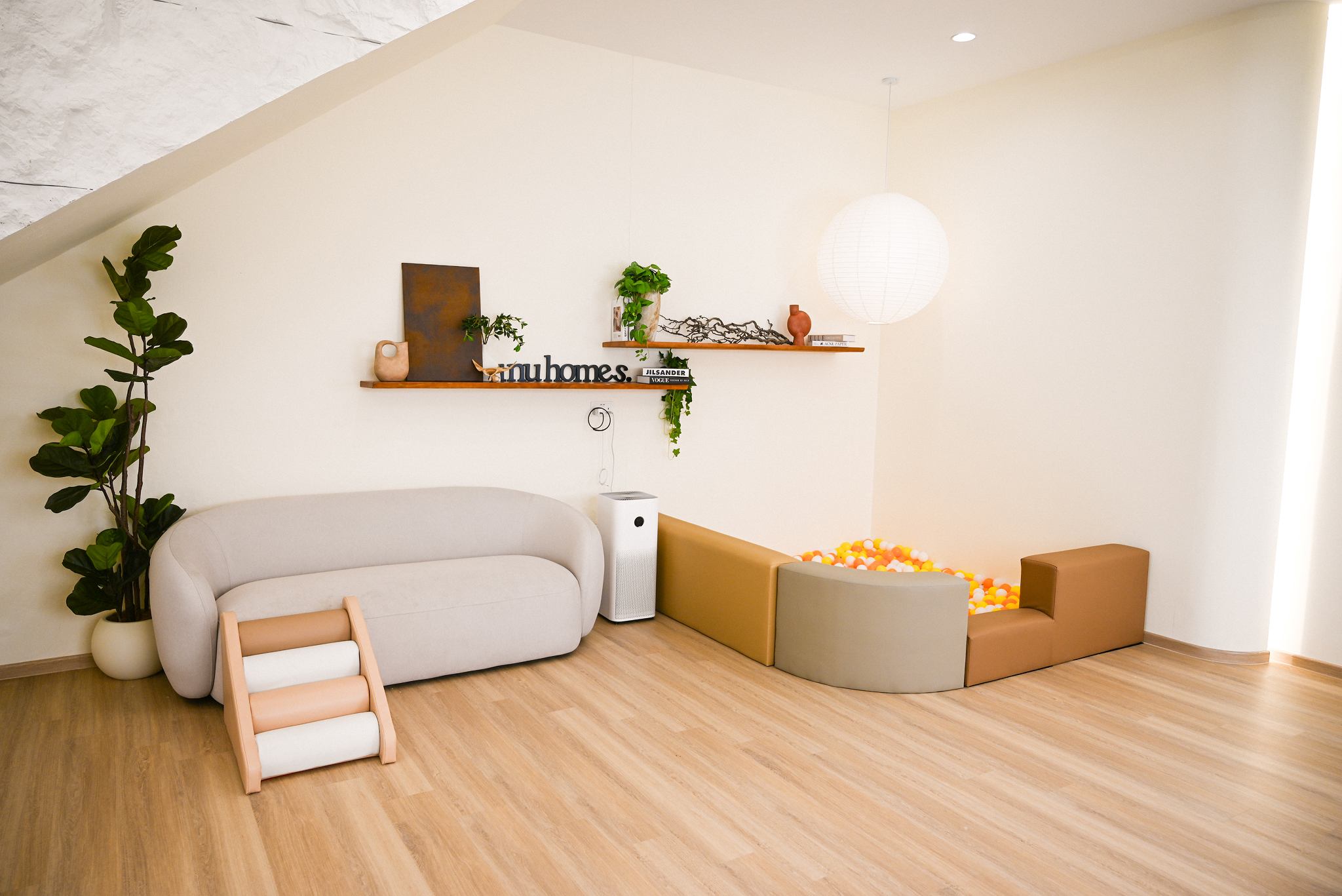 Modern minimalist room with a light gray sofa, indoor plant, floating shelves with decor, white air purifier, and a ball pit with orange, yellow, and white balls under a white hanging paper lantern.