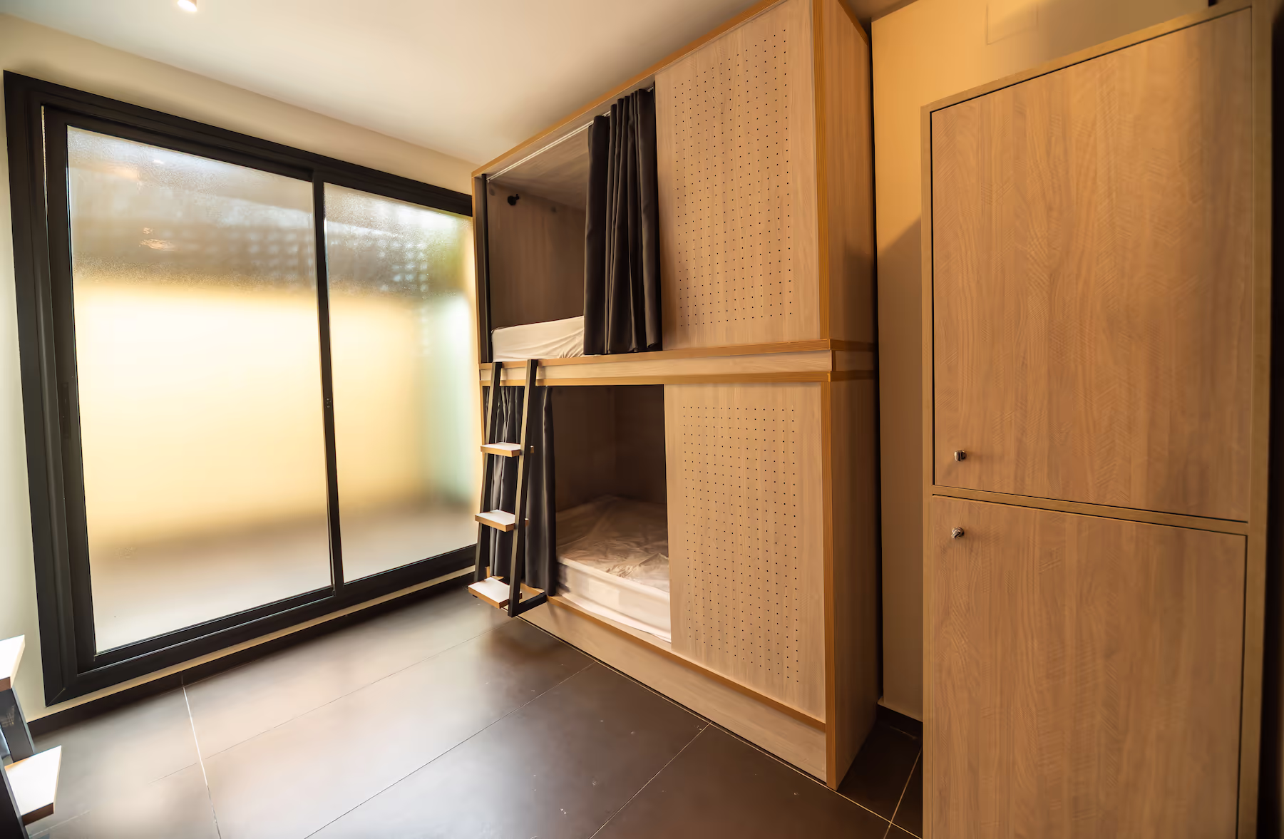 Modern room with wooden bunk beds featuring black privacy curtains and a small ladder, next to a wooden cabinet and a large frosted glass sliding door.