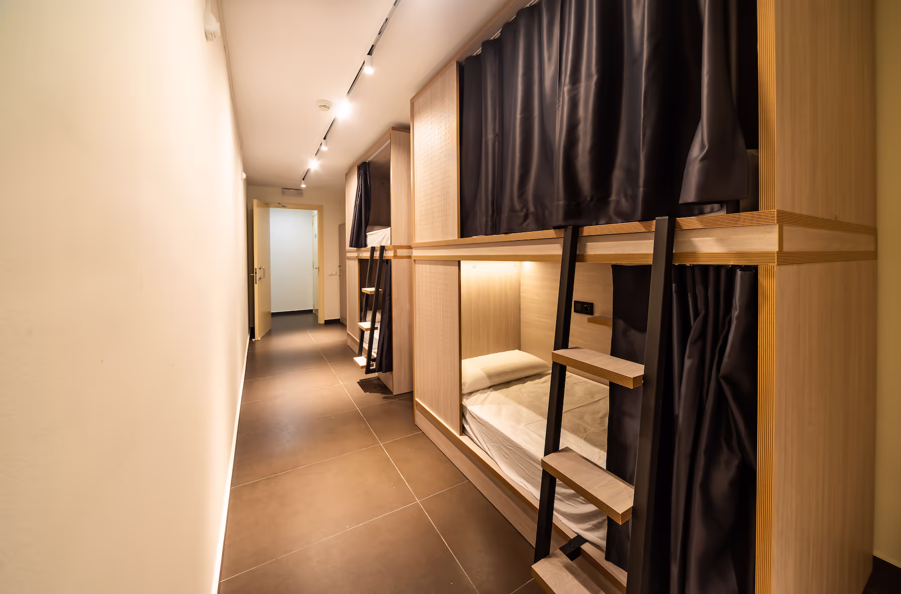 Narrow hostel room with wooden double-decker bunk beds featuring black privacy curtains and small ladders.