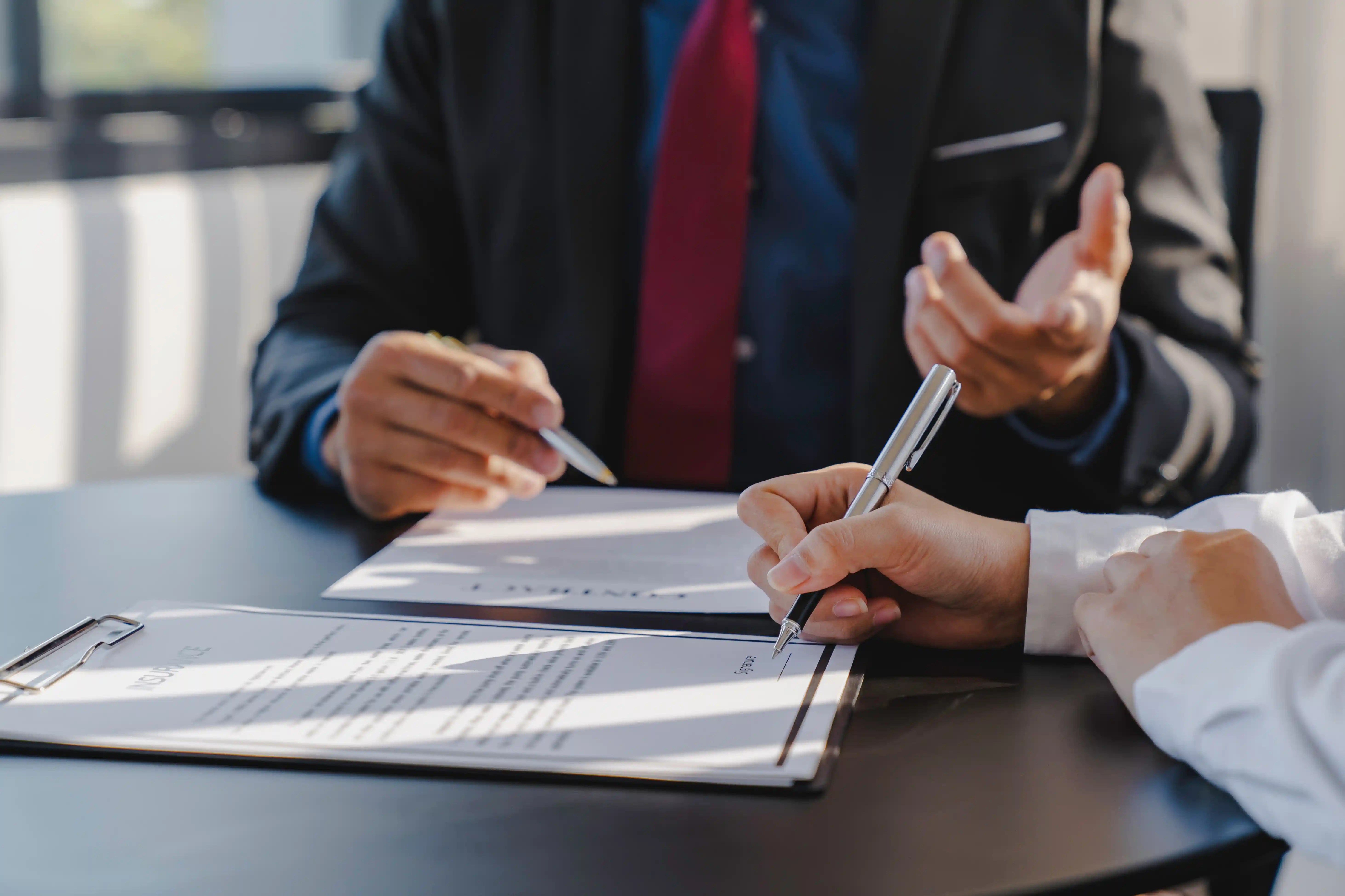 Person signing a document on a clipboard at a table while another person in a suit gestures and holds a pen.