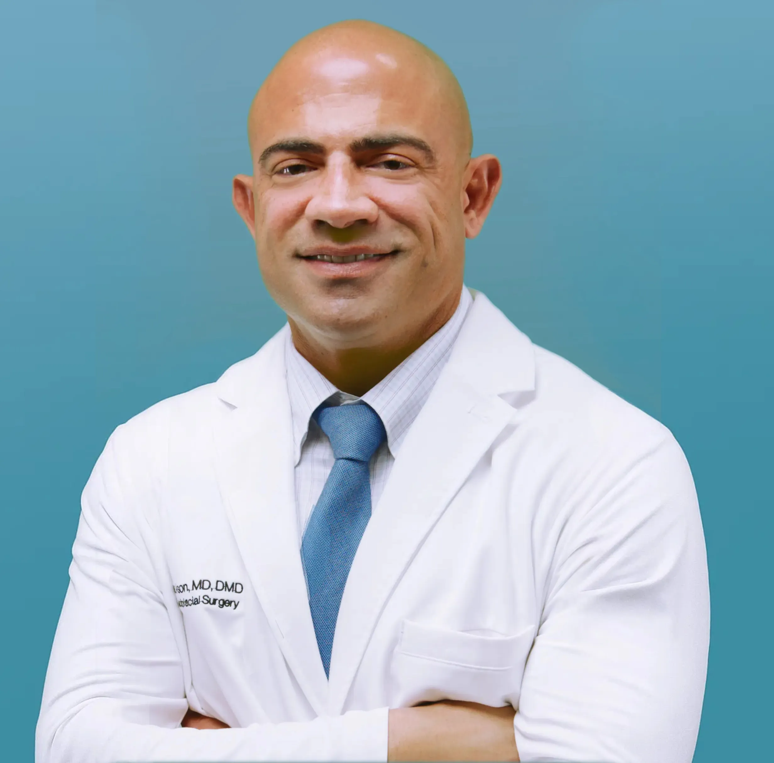 Dr Ason wearing a white medical coat and blue tie, standing with arms crossed against a blue background.