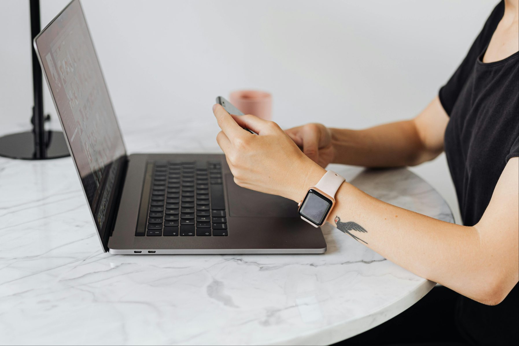 Personne portant une montre connectée avec un tatouage d’oiseau sur l’avant-bras, utilisant un smartphone à côté d’un ordinateur portable sur une table en marbre