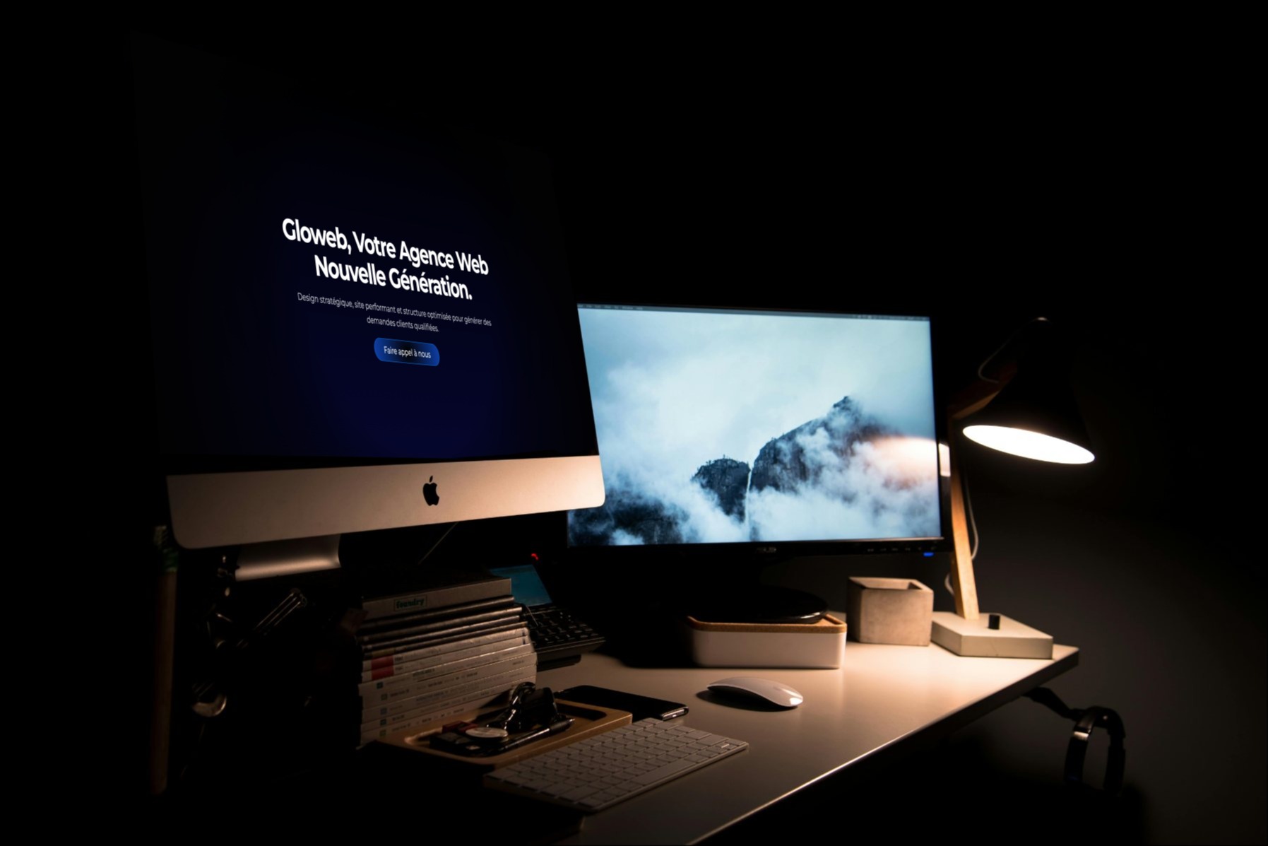 Bureau avec deux écrans d’ordinateur, l’un affichant un site d’agence web en français et l’autre un paysage de montagne brumeux, éclairés par une lampe de bureau.