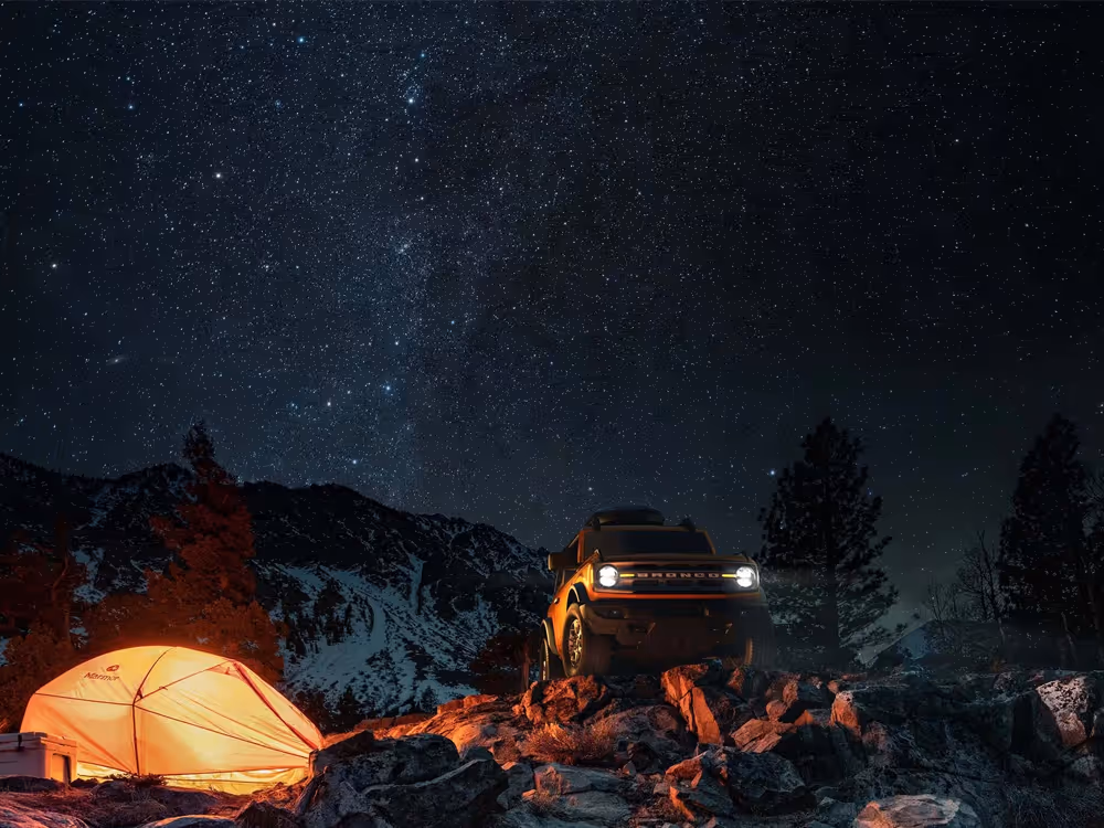 Ford Bronco parked beside glowing tent under starry night sky in mountain landscape, cinematic adventure photography by Roe Photo.