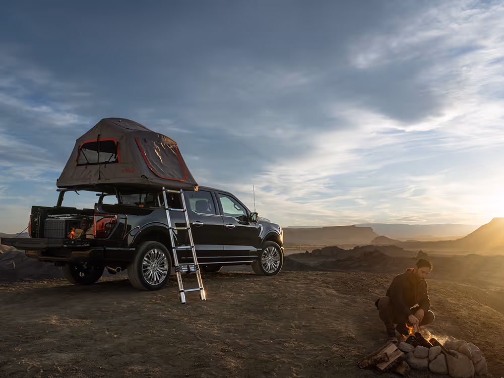 Adventurer by campfire near black pickup truck with rooftop tent at golden hour, outdoor lifestyle photography by Roe Photo.