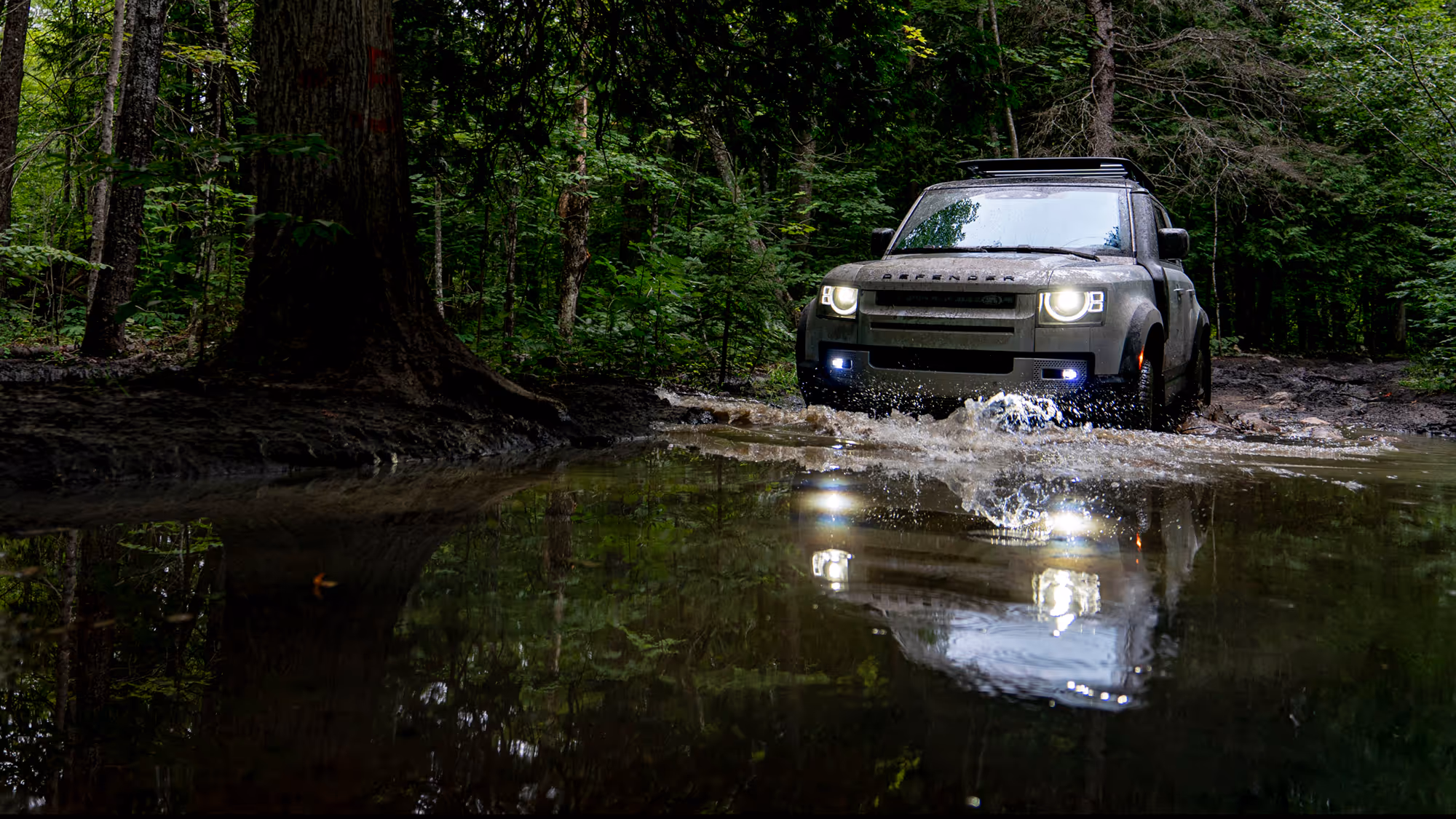 A rugged vehicle driving through a muddy area in a forest, splashing water. Sunlight filters through the trees, reflecting off the water surface. This photography highlights the intersection of nature and technology in Metro Detroit.