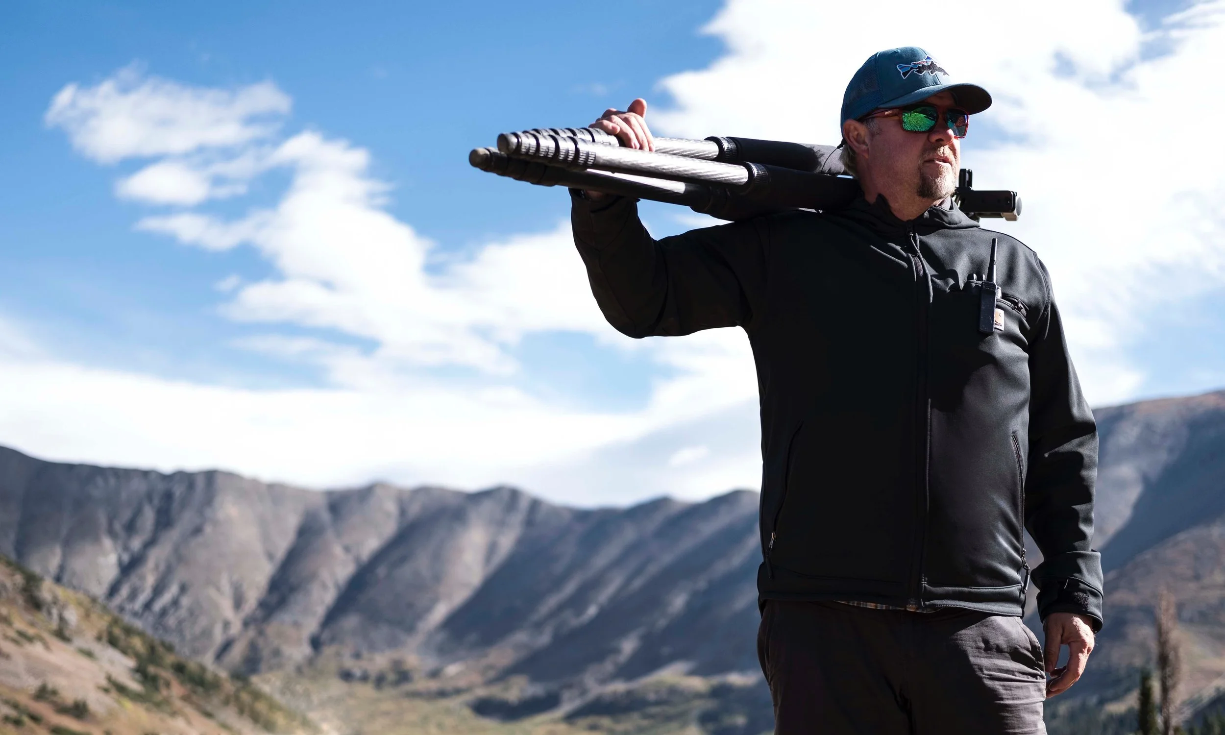 A man wearing sunglasses and a cap carries a tripod over his shoulder while standing in a mountainous landscape. The background features rugged mountains and a blue sky with clouds, illustrating a connection between Photography, Metro Detroit, Technology, AI, and Artistry.