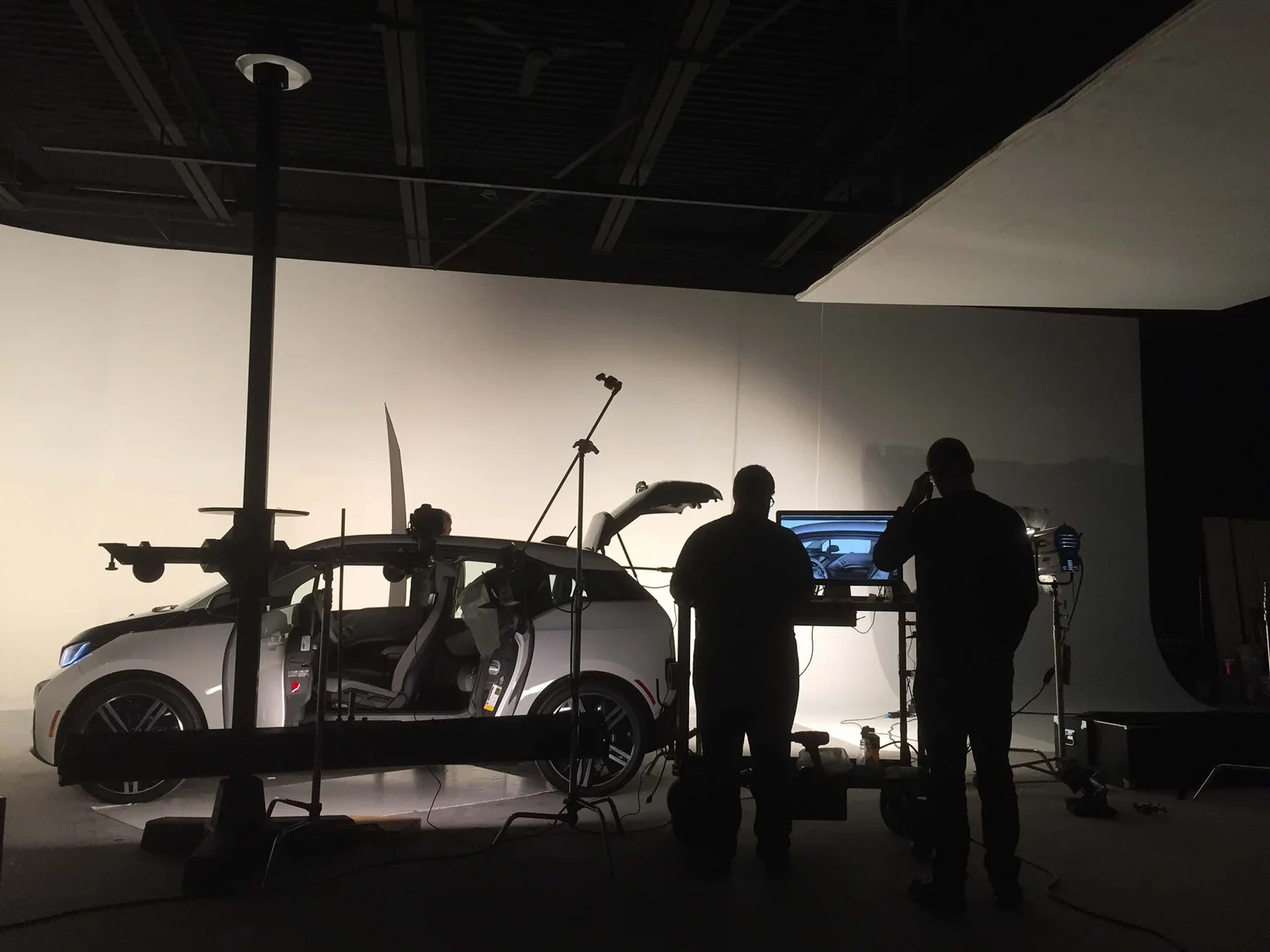 A behind-the-scenes view of a photography studio featuring a car with its doors open. Two silhouetted figures operate equipment in front of a screen, capturing the car's image. The setting highlights the integration of technology and artistry in Metro Detroit.