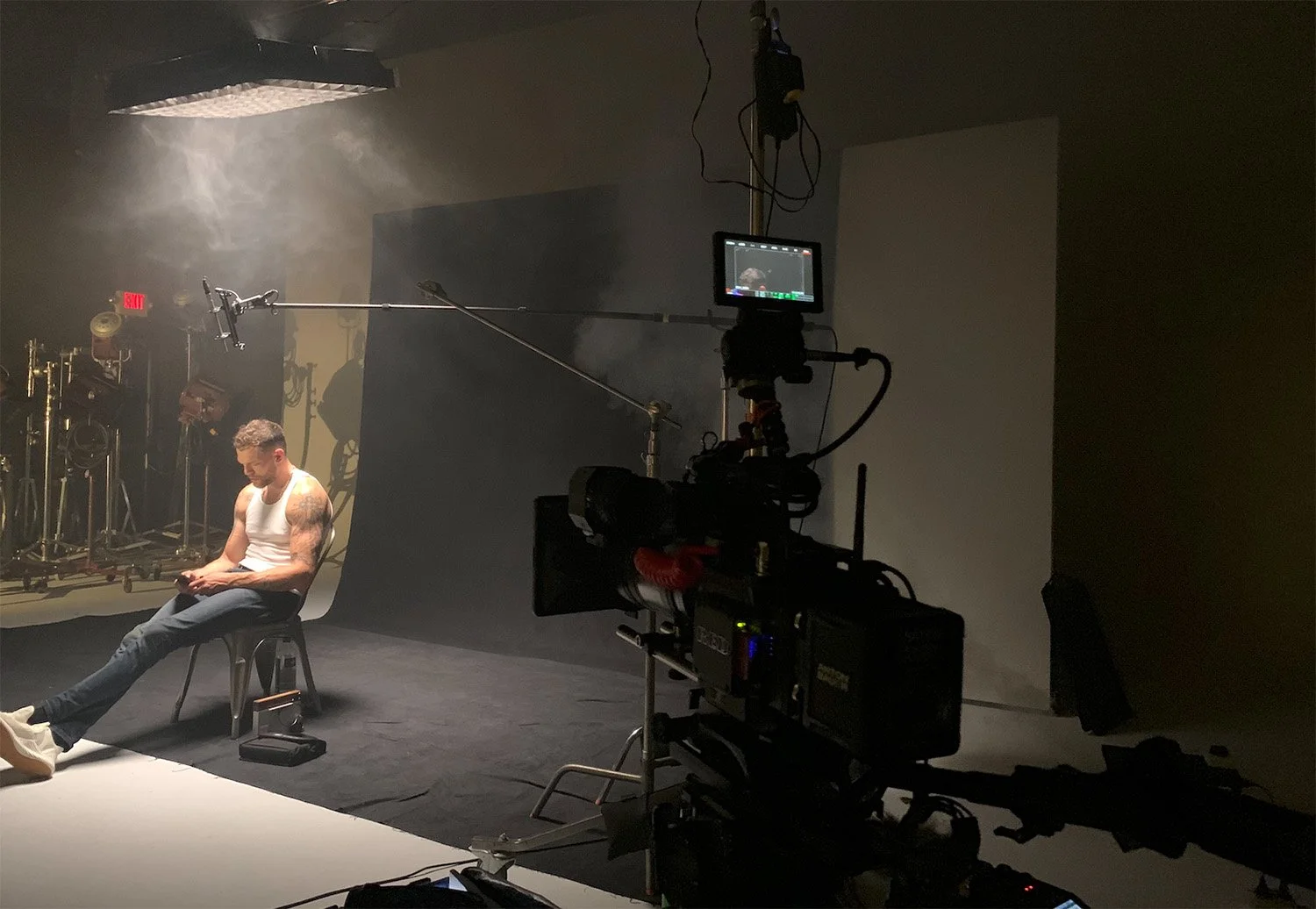 A man sitting on a chair in a photography studio, illuminated by a soft light. A camera setup with visible equipment captures the scene, surrounded by a misty atmosphere. Keywords include Photography, Metro Detroit, Technology, AI, and Artistry.