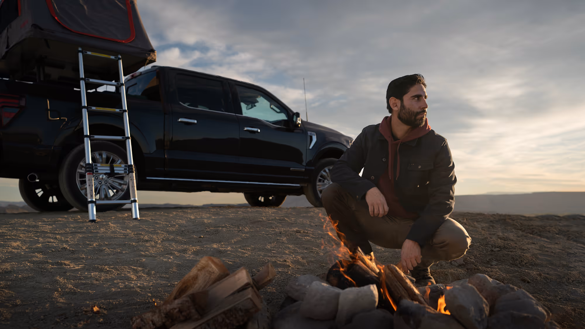 Man sitting near campfire beside black pickup truck with rooftop tent at sunset, outdoor adventure lifestyle photo by Roe Photo.