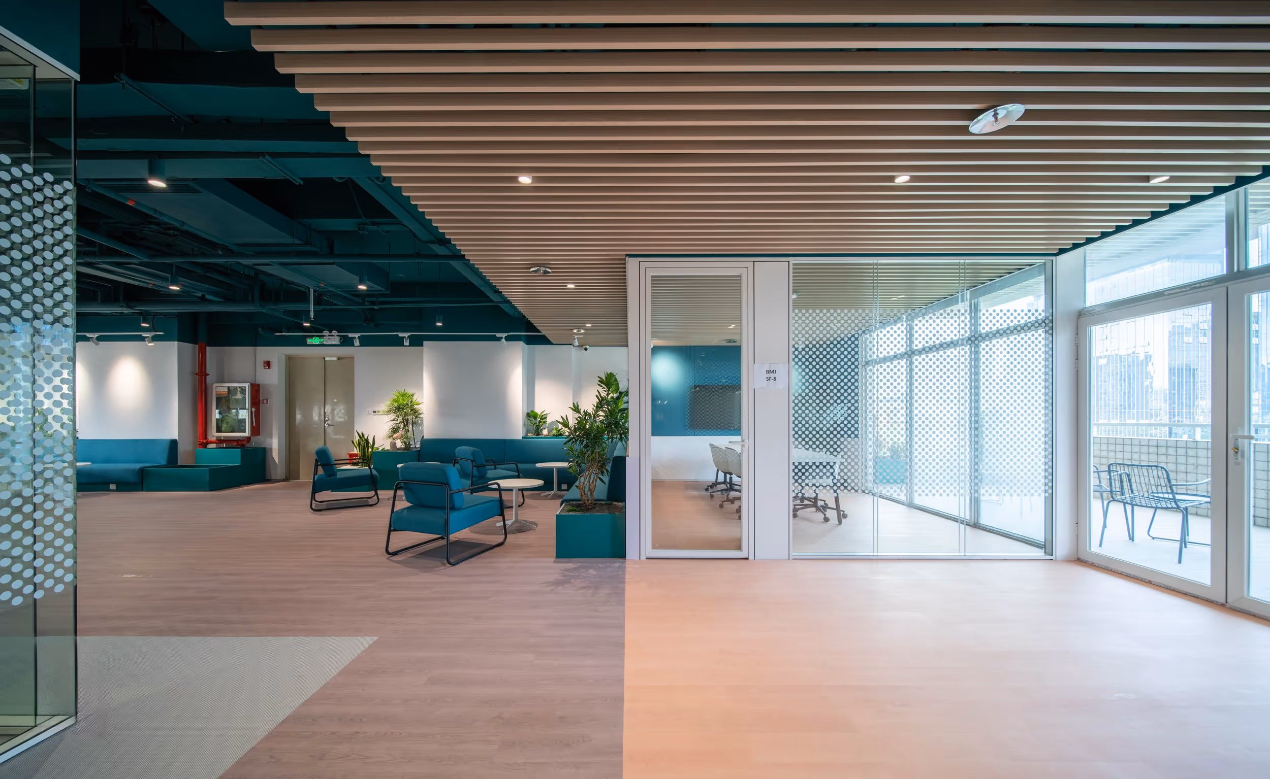 Modern office lounge area with teal chairs, potted plants, and a glass-walled conference room next to a balcony with city views.