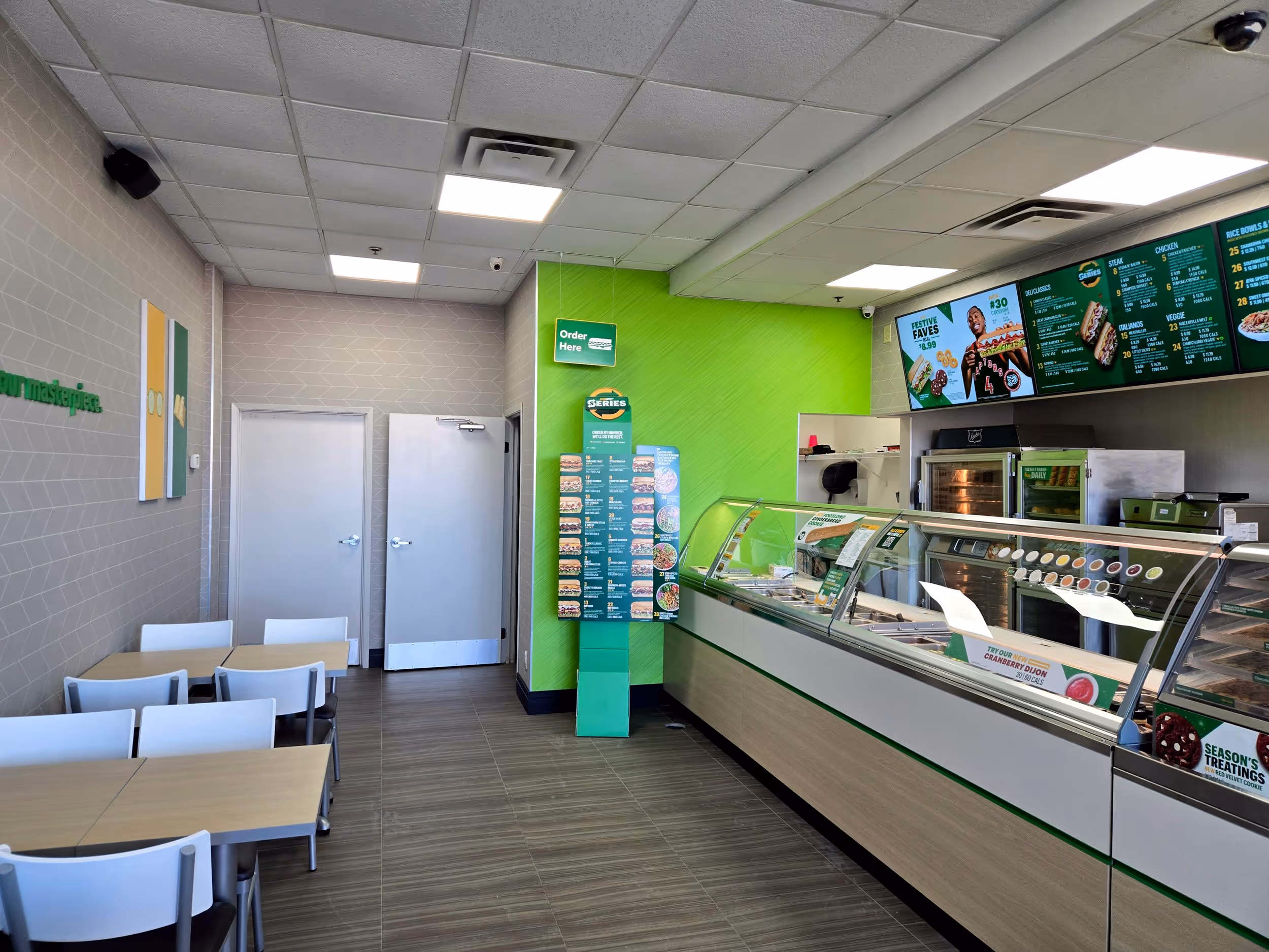 Interior of a brightly lit sandwich shop with a green accent wall, digital menu screens, glass food display counter, and seating area with tables and chairs.